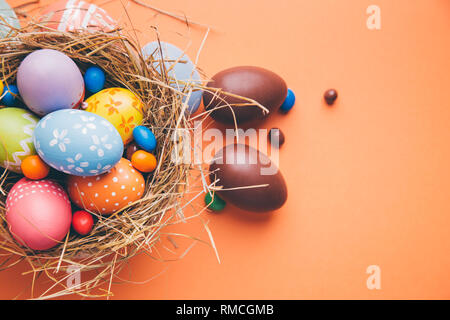 Easter eggs painted by children with chocolate and candies in a hay nest on a orange background. Top view Stock Photo