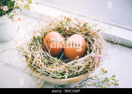 Easter eggs with rubber stamp "Frohe Ostern" greeting text in a hay and flowers nest next to a window at home Stock Photo