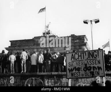 A crowd is standing on the Berlin Wall looking towards the Brandenburg Gate. In the foreground a sign with the inscription 'Attention, you are now leaving West Berlin'. This was overwritten with the grafitti 'How'. Stock Photo