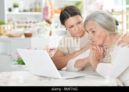 Portrait of sad mother and daughter sitting at table with laptop at home Stock Photo