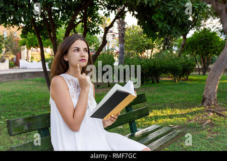 Young woman is sitting on the bench in the park and thinking about what she has read. Stock Photo
