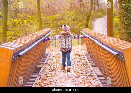 Girl in a coat and hat in an autumn day walks on a wooden bridge. A girl walks across the bridge on an autumn day. An autumn warm sunny day. Stock Photo