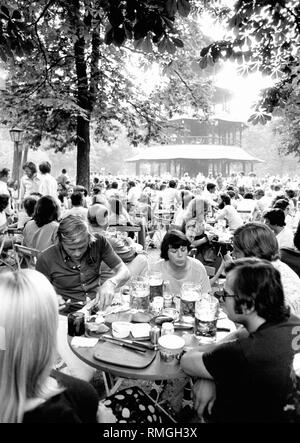 Many people sit in the beer garden at the Chinese Tower in the Englischer Garten in Munich. Here is also a table with students (among them the future lawyer Peter Steinmaier and the economist Hans Stettner), who are taking a light meal. Stock Photo