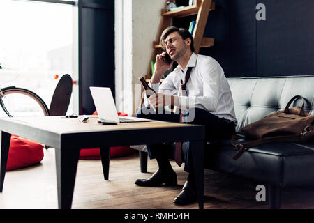 tired man in suit holding bottle while talking on smartphone after party Stock Photo