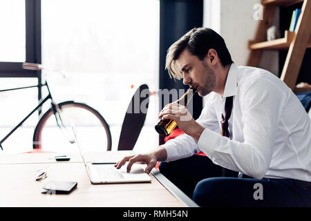 tired man in suit drinking beer from bottle while using laptop after party Stock Photo