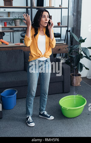 Full length view of shocked woman talking on phone while water leaking from ceiling Stock Photo