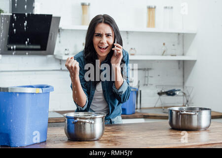 Irritated woman calling plumber while water leaking in bucket from ceiling Stock Photo