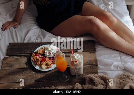 Young Woman eating appetizing waffles and tasty drinks on tray on bed Stock Photo