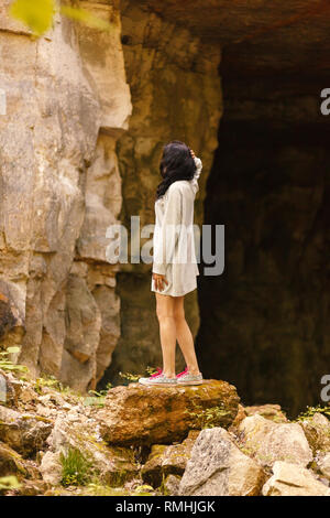 A woman in a long t-shirt and sneakers with pink laces stands on a large stone on the background of high rocks. Woman standing in front of cave entran