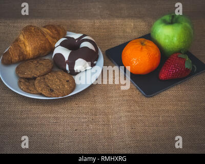 A plate of black slate with a green apple, a tangerine and a strawberry and one plate with one croissant, one chocolate donut and cookies on a burlap  Stock Photo