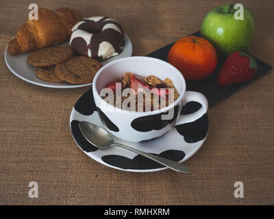 A bowl with cornflakes and red berries and a plate of black slate with a green apple, a tangerine and a strawberry and one plate with one croissant, o Stock Photo
