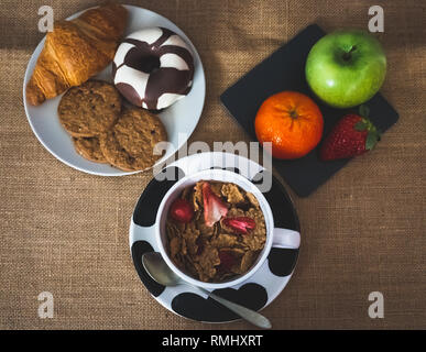 A bowl with cornflakes and red berries and a plate of black slate with a green apple, a tangerine and a strawberry and one plate with one croissant, o Stock Photo