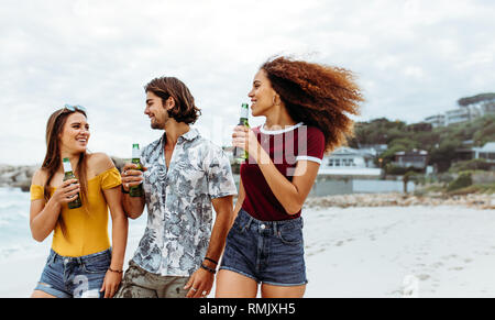 Group of three young friends walking outdoors with beer bottles. Multi-ethnic friends with beers walking along the beach. Stock Photo