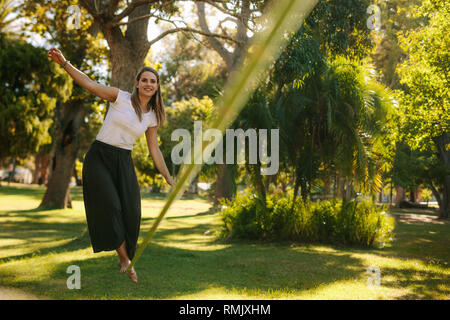 Woman balancing her walk on a loose rope tied between two trees. Woman practicing slack rope walking in a park. Stock Photo
