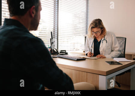 Female doctor making notes in her book while consulting a patient. Doctor writing medical history of male patient sitting across her desk in hospital. Stock Photo