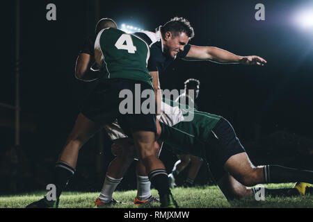 Rugby players making an effort to get the ball from opponent team player. Rugby players competing in match under lights. Stock Photo