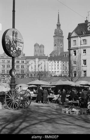 View of the Munich Viktualienmarkt. On the left next to the lamppost, a promotional sign with the inscription: 'Muenchner Qualitaets-Gemuese taeglich frisch geerntet' ('Quality fresh vegetables harvested daily in Munich'). In the background, the church towers of the Frauenkirche and of the St. Peter. Stock Photo