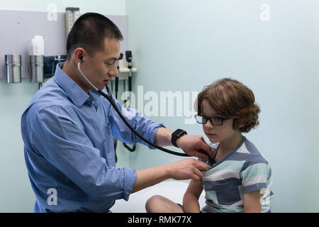 Young asian male doctor examining caucasian boy patient with stethoscope in clinic Stock Photo