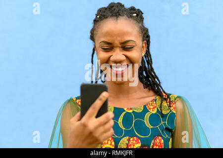 Happy cute young African girl in a stylish dress reading a text message on her mobile phone and pulling a funny face isolated on blue Stock Photo