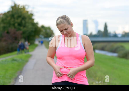 Portrait of an upset young woman looking annoyed at her belly fat while wearing fitness pink sleeveless top outdoors Stock Photo