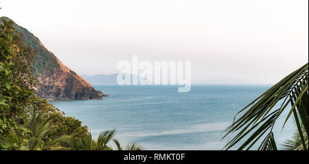 Beautiful Landscape clicked after getting lost while vacating on remote island. Photo comprise calm blue sea water, hazy horizon, mountains, palm leaf Stock Photo