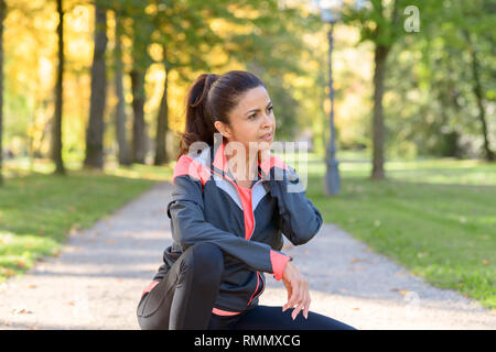 Athletic woman taking a break from a workout sitting in a road through a park rubbing her neck Stock Photo
