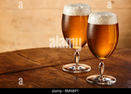 Two tall stemmed glasses of fresh chilled draft or craft beer with fresh frothy heads standing side by side on an old table with lateral copy space Stock Photo