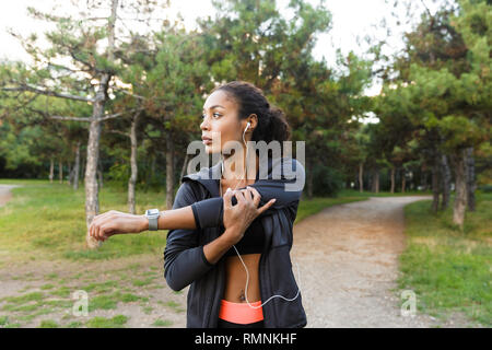 Portrait of energetic african american woman 20s wearing black tracksuit doing exercises and stretching her body in green park Stock Photo