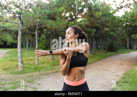 Young african american woman 20s wearing black tracksuit doing exercises and stretching her body in green park Stock Photo
