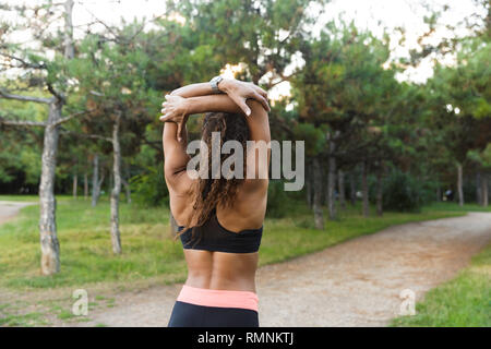Beautiful african american woman 20s wearing black tracksuit doing exercises and stretching her body in green park Stock Photo