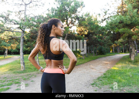 Slim african american woman 20s wearing black tracksuit doing exercises and stretching her body in green park Stock Photo