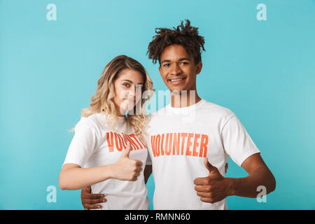 Happy confident multiethnic couple wearing volunteers t-shirt standing isolated over blue background, thumbs up Stock Photo