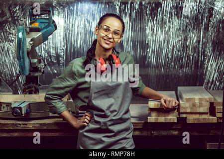 Smiling craftswoman grinding timbers with special machine. Beautiful woman wearing safety glasses. Concept of joiner's shop and woodworking. Gender equality. Male profession Stock Photo