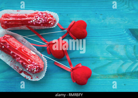 couple of red raspberry eclairs with red velvet hearts on blue wooden background with space for text Stock Photo