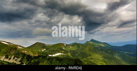 Wide panorama of lit by morning sun green valley, hills covered with forest and distant misty mountains with patches of snow under cloudy windy sky. B Stock Photo