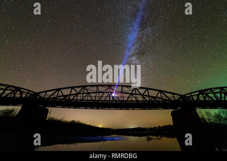 Man with flashlight standing on river bank under illuminated metal bridge under dark starry sky reflected in water. Night photography concept. Stock Photo