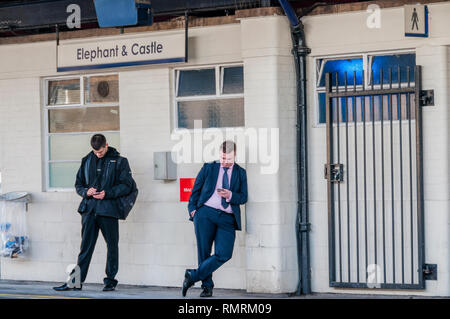 Two passengers looking at their mobile phones on the platform at Elephant & Castle station in south London, while waiting for a train. Stock Photo