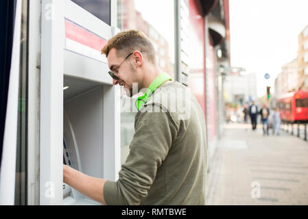 Young handsome man with sunglasses and headphones taking money from the ATM Stock Photo