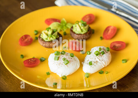 Poached eggs with avocado stuffed mushrooms, tomatoes, and green onions on a cheery yellow plate on a wooden table Stock Photo