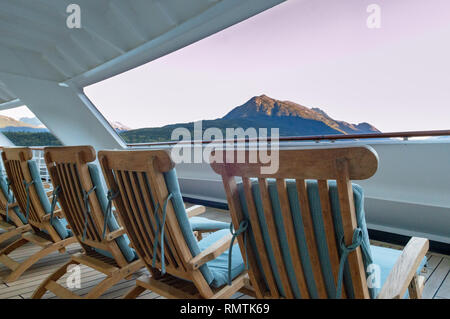 Row of open cushioned wooded deck lounge chairs at sunrise in the early morning on outdoor veranda at stern of cruise ship, Skagway, Alaska, USA. Stock Photo
