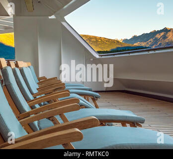 Row of open cushioned wooded deck lounge chairs at sunrise in the early morning on outdoor veranda at stern of cruise ship, Skagway, Alaska, USA. Stock Photo