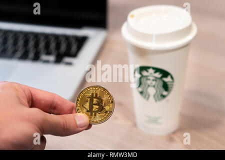 Cryptocurrency Bitcoin held by a humand hand next to Starbucks coffee cup. Slovenia 13.02.2019 Stock Photo