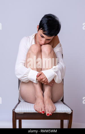 Lonely depressed woman sitting on a chair resting her chin on her bent knees as she stares disconsolately down at the floor Stock Photo