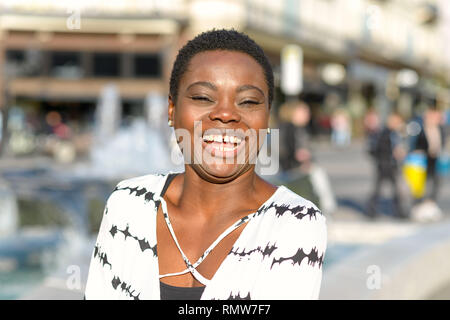 Happy vivacious young African woman in the city standing in the sunshine in the street laughing at the camera Stock Photo