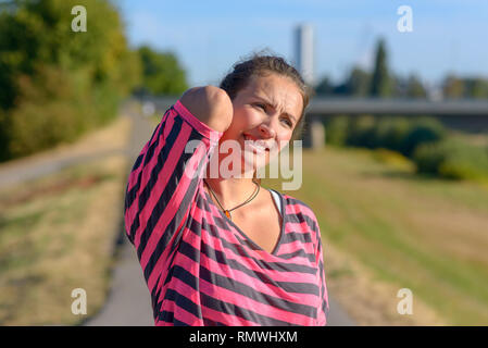 Young woman out jogging suffers a muscle injury standing holding her neck and lower back while grimacing in pain on a rural road, close up upper body  Stock Photo