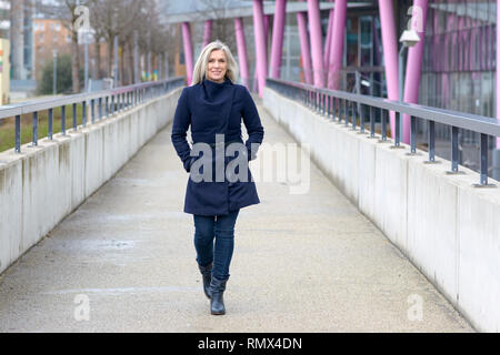 Trendy blond woman in a stylish blue outfit walking along a walkway in town approaching the camera with a friendly smile Stock Photo