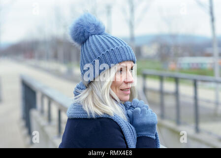Pretty blond woman cuddling down into her warm knitted blue woolly scarf, hat and gloves as she stands waiting outdoors on a walkway in the city Stock Photo