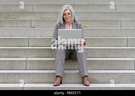 Middle-aged businesswoman using her laptop in town sitting on a flight of outdoor steps smiling as she browses the internet Stock Photo