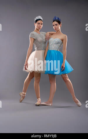 Young attractive ballet dancers standing together in studio, looking at camera and posing. Two ballerinas wearing blue and beige dresses, ballet shoes and crowns standing on socks. Concept of ballet. Stock Photo