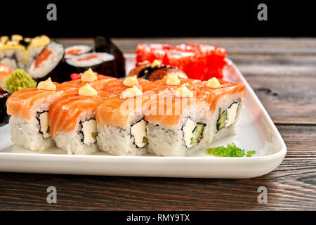 Close up of delicious philly rolls, covered with salmon and decorated with mayo. Sushi set of different kinds of uramaki and maki rolls, served on white plate. Stock Photo